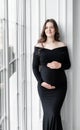 beautiful young pregnant woman in dark black dress stands near window Royalty Free Stock Photo