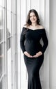 beautiful young pregnant woman in dark black dress stands near window Royalty Free Stock Photo