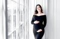 beautiful young pregnant woman in dark black dress stands near window Royalty Free Stock Photo