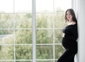 beautiful young pregnant woman in dark black dress stands near window Royalty Free Stock Photo