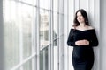 beautiful young pregnant woman in dark black dress stands near window Royalty Free Stock Photo