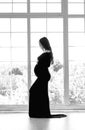 beautiful young pregnant woman in dark black dress stands near the window black and white photo Royalty Free Stock Photo