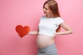 Happy pregnant woman holding red heart near the belly, on a pink background Royalty Free Stock Photo