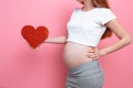 Happy pregnant woman holding red heart near the belly, on a pink background Royalty Free Stock Photo