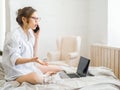 Beautiful young pregnant woman sitting on bed using technology devices: laptop and phone in modern room. Future mom Royalty Free Stock Photo