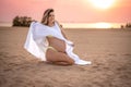 Beautiful young pregnant woman sitting on sand, holding her white dress and enjoying pregnancy on the beach at sunset. Royalty Free Stock Photo