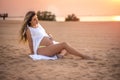 Beautiful young pregnant woman sitting on sand, touching her belly and enjoying pregnancy on the beach at sunset. Royalty Free Stock Photo
