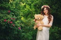 Beautiful young pregnant woman wearing white dress and floral wreath and holding teddy bear in the summer green park, summertime, Royalty Free Stock Photo