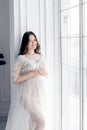 beautiful young pregnant woman in white dress stands near window Royalty Free Stock Photo