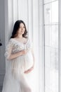 beautiful young pregnant woman in white dress stands near window Royalty Free Stock Photo