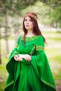 A beautiful, young, red-haired girl in a green medieval dress with long sleeves, with a golden crown, standing on the grass, in a Royalty Free Stock Photo