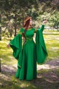 A beautiful, young, red-haired girl in a green medieval dress with long sleeves, with a golden crown, standing on the grass, in a Royalty Free Stock Photo