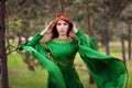 A beautiful, young, red-haired girl in a green medieval dress with long sleeves, with a golden crown, standing on the grass, in a Royalty Free Stock Photo