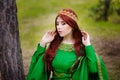 A beautiful, young, red-haired girl in a green medieval dress with long sleeves, with a golden crown, standing on the grass, in a Royalty Free Stock Photo