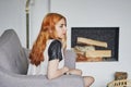 Beautiful young red-haired girl sitting on the sofa in the living room against the background of the fireplace Royalty Free Stock Photo