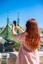 Beautiful young red-haired girl in a summer Cotton dress photographs a landmark bridge on a sunny summer day. Vertical photo Royalty Free Stock Photo