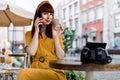 Beautiful young red haired girl wearing stylish yellow suit, talking on smart phone in a city cafe outdoors, looking at Royalty Free Stock Photo