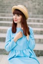 Beautiful young red haired woman wearing blue dress and hat, posing to camera while sitting on city stairs outdoors Royalty Free Stock Photo