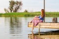 Beautiful young red woman sitting on a wooden pier in a colorful long sarafan dress and putting her leg in a water Royalty Free Stock Photo