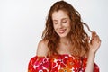 Beautiful young redhead woman playing with her curly hair, looking down with flirty coquettish smile, standing against Royalty Free Stock Photo