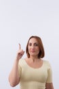 Beautiful young redheaded woman looking at camera smiling and pointing finger upward on light background. Royalty Free Stock Photo