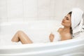 Beautiful young relaxed brunette woman wearing white towel on her head enjoying her bath with bubbles in bright bathroom Royalty Free Stock Photo