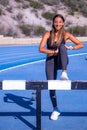 Beautiful young runner girl, tanned with long brown hair, smiling with her leg bent leaning on an obstacle on a running track Royalty Free Stock Photo