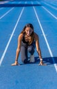 Beautiful young runner girl, tanned with long dark hair, smiling looking at the track at the starting line of a blue running track Royalty Free Stock Photo