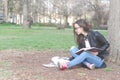 Beautiful young school or college girl with long hair, eyeglasses and black leather jacket sitting on the ground in the park readi Royalty Free Stock Photo