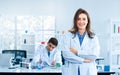 A beautiful young scientist stands smiling in a science lab with a male scientist sitting in the back Royalty Free Stock Photo