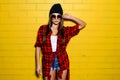 Beautiful young girl posing and smiling near yellow wall background in sunglasses, red plaid shirt, shorts, hat. Royalty Free Stock Photo