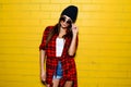 Beautiful young girl posing and smiling near yellow wall background in sunglasses, red plaid shirt, shorts, hat. Royalty Free Stock Photo