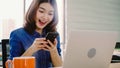 Beautiful young smiling asian woman working on laptop while enjoying using smartphone at office Royalty Free Stock Photo