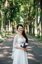 Beautiful young smiling brunette bride in wedd dress with bouquet of flowers in hands outdoors on the background of green leaves Royalty Free Stock Photo