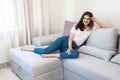 Beautiful young smiling brunette woman wearing jeans and white t-shirt sitting on the sofa in bright livingroom looking happy