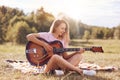 Beautiful young smiling female plays acoustic guitar, sits cross legs on green grass, being in good mood, learns new song, has app Royalty Free Stock Photo