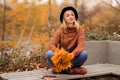 Beautiful young smiling girl blonde in a brown warm sweater, black felt hat, blue jeans and boots sits on a bench, cross-legged, Royalty Free Stock Photo