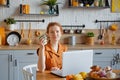 A beautiful young smiling red-haired woman is working on a laptop and drinking coffee while sitting at a table in the kitchen at Royalty Free Stock Photo