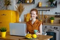 A beautiful young smiling red-haired woman is working on a laptop and drinking coffee while sitting at a table in the kitchen at Royalty Free Stock Photo