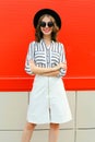 Beautiful young smiling woman crossed her arms wearing white striped shirt, skirt, black round hat on city street over red wall Royalty Free Stock Photo