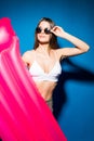 Beautiful young smiling woman dressed in white swimsuit and glasses posing with pink inflatable mattress isolated over blue backgr Royalty Free Stock Photo