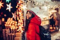 Beautiful young smiling woman enjoy snow winter time on Christmas fair in night city wearing hat and red jacket. Royalty Free Stock Photo