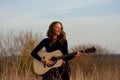 beautiful young smiling woman playing an acoustic guitar while standing in field among tall dried grass. Royalty Free Stock Photo