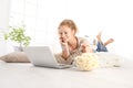 beautiful young smiling woman watch a movie at the computer eating popcorn lying on living room wooden floor in comfortable home Royalty Free Stock Photo