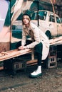 Beautiful young stylish blonde woman wearing long beige coat, white boots, black hat posing through the city streets. Trendy