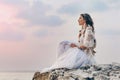 beautiful young stylish boho woman sitting on the beach at sunset