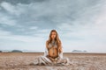 Beautiful young stylish pregnant woman with boho accessories sitting on the beach at sunset