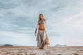 Beautiful young stylish pregnant woman with boho accessories walking on the beach at sunset