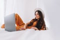 Beautiful young stylish woman in white hat lying on bed, using laptop computer Royalty Free Stock Photo