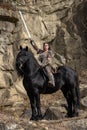 Beautiful young viking warrior woman with sword and big black horse in the mountains scouting the horizont